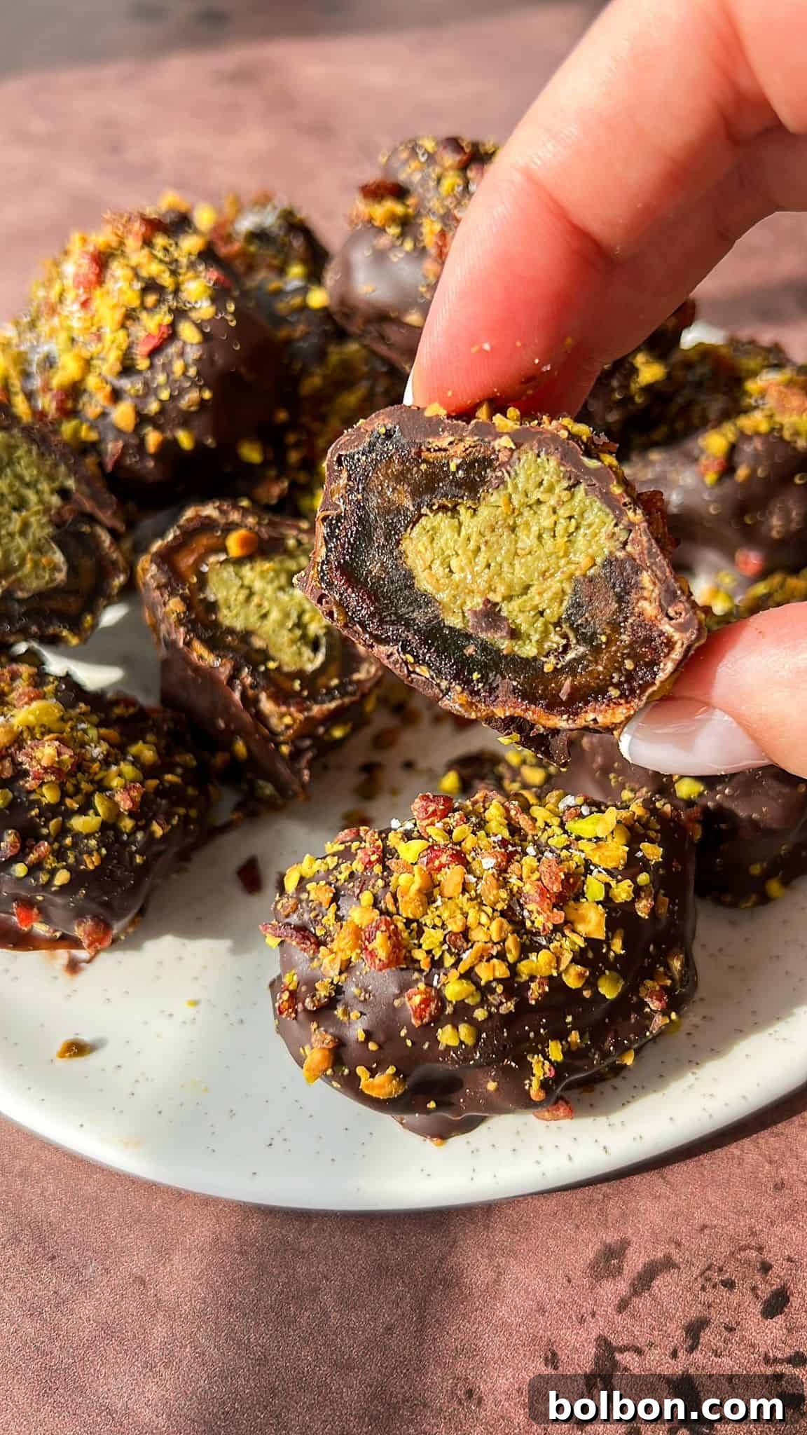 Exquisite Dubai Chocolate Dates 2 A hand holds a half-eaten chocolate-covered pistachio ball over a plate. The treat is coated in dark chocolate and sprinkled with crushed pistachios. Other similar treats are visible on the plate.