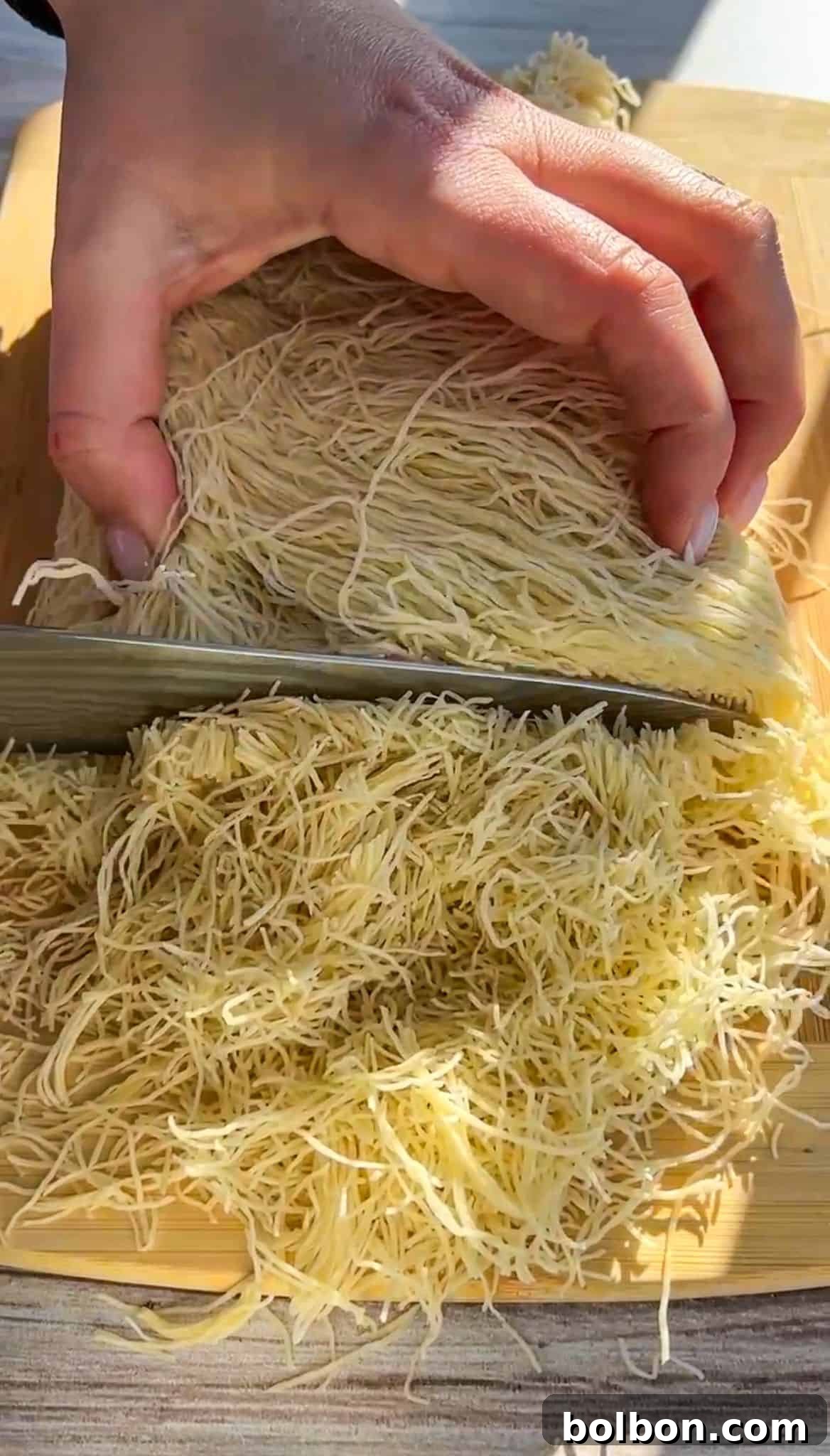 Exquisite Dubai Chocolate Dates 3 A hand holding a knife slices through a block of fine shredded dough, possibly kataifi, on a wooden cutting board placed on a light-colored table. The dough is spread out as it's being cut.