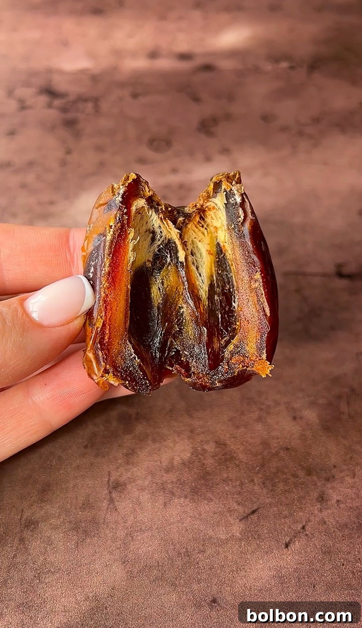 Exquisite Dubai Chocolate Dates 8 A hand with a manicured nail holds a halved date, revealing its sticky, fibrous interior. The background is a soft, brown surface.