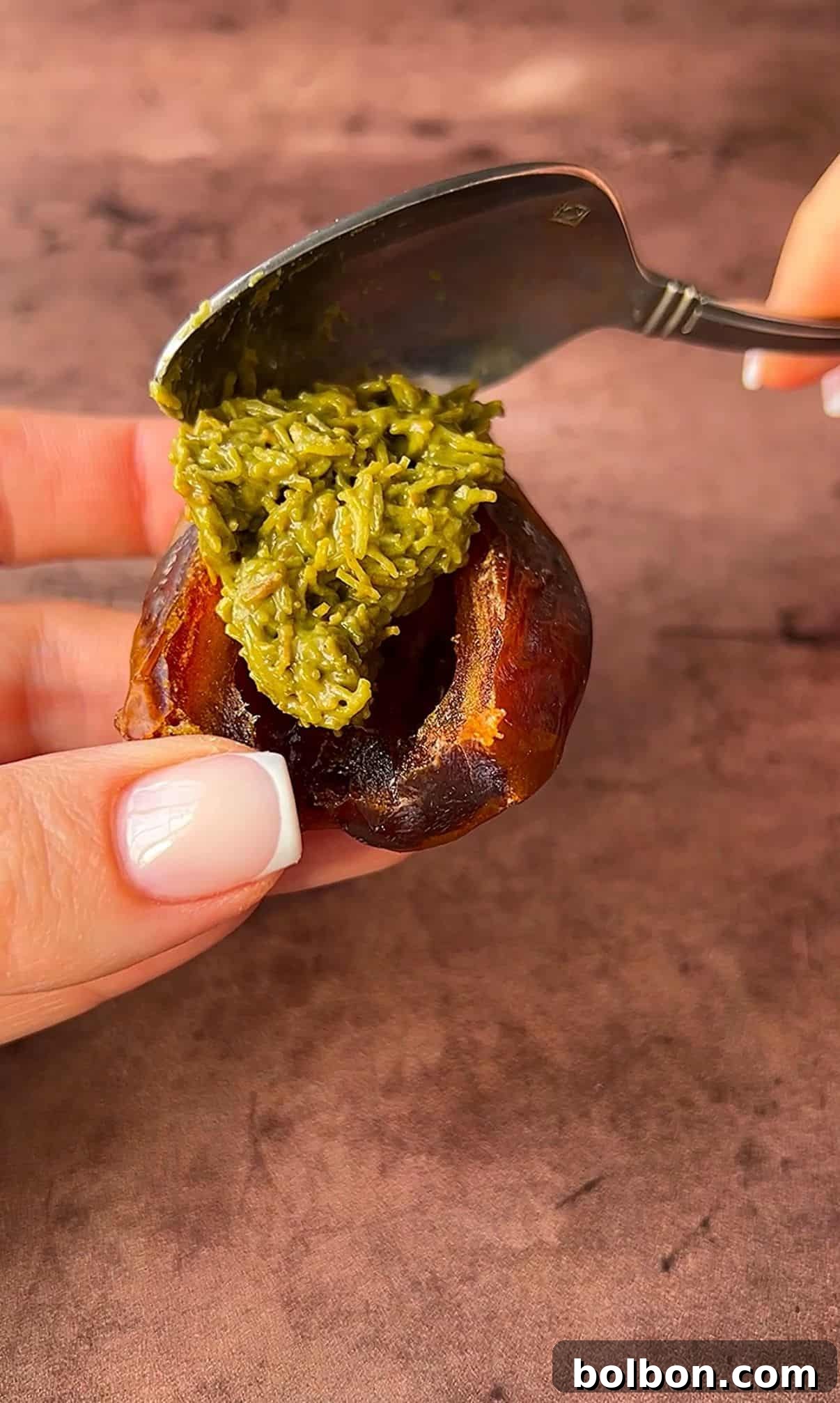 Exquisite Dubai Chocolate Dates 9 A hand is holding a date, and another hand is using a spoon to fill it with a green paste. The background is a brown surface.