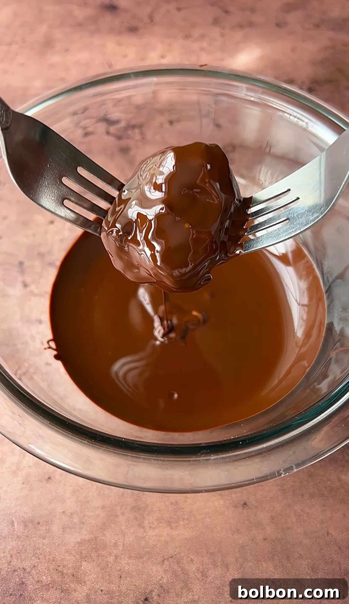 Exquisite Dubai Chocolate Dates 10 A Dubai chocolate date is held between two forks above a glass bowl filled with more melted chocolate. The background is a textured brown surface.