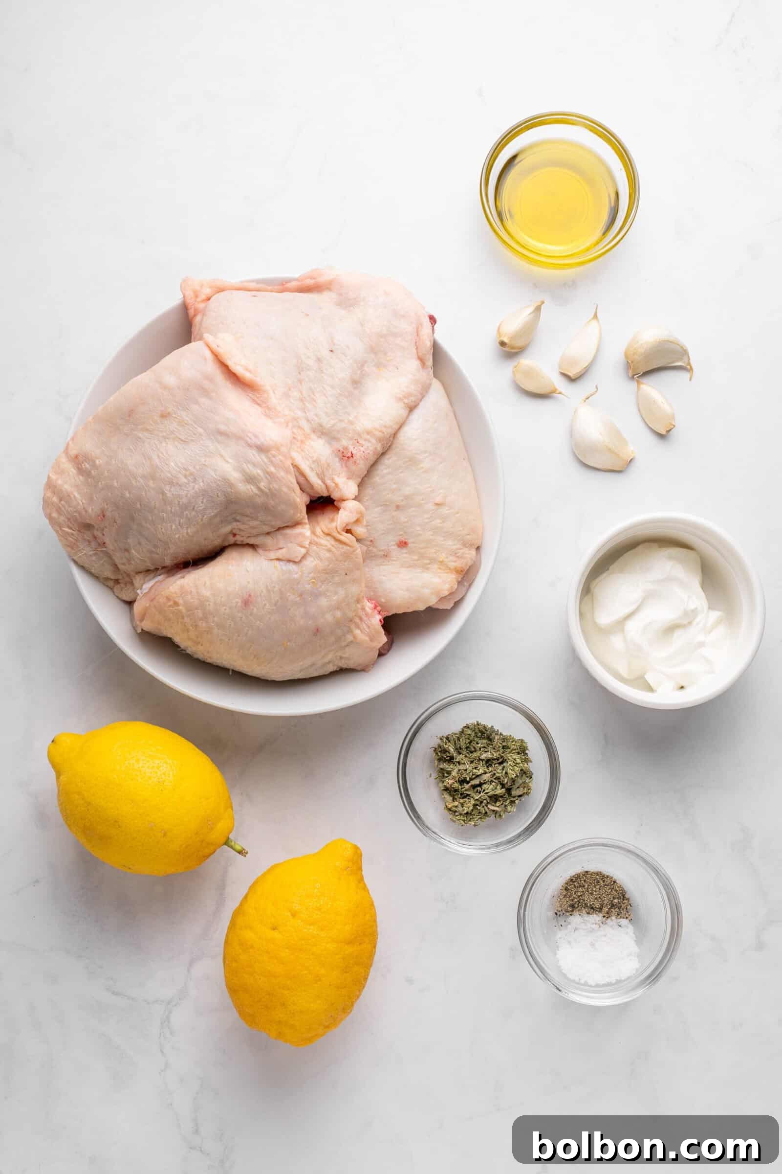 Zesty Lemon Yogurt Chicken 3 A flat lay of raw chicken thighs in a bowl, surrounded by garlic cloves, a small bowl of olive oil, two lemons, a bowl of yogurt, herb seasoning, salt, and pepper, all on a white marble surface.
