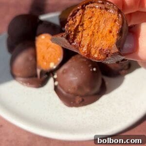 A hand holds up a chocolate-covered dessert with a bite taken, revealing a caramel or nougat-like filling. More of these treats are piled on a white plate in the background, all on a brown table surface.