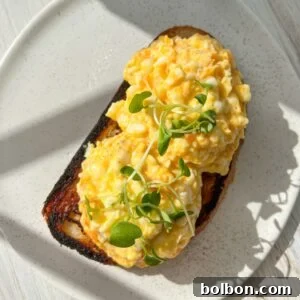 A plate with two slices of toast topped with scrambled eggs and garnished with microgreens. The toast is slightly charred, and sunlight casts shadows on the white, speckled plate and wooden table surface.
