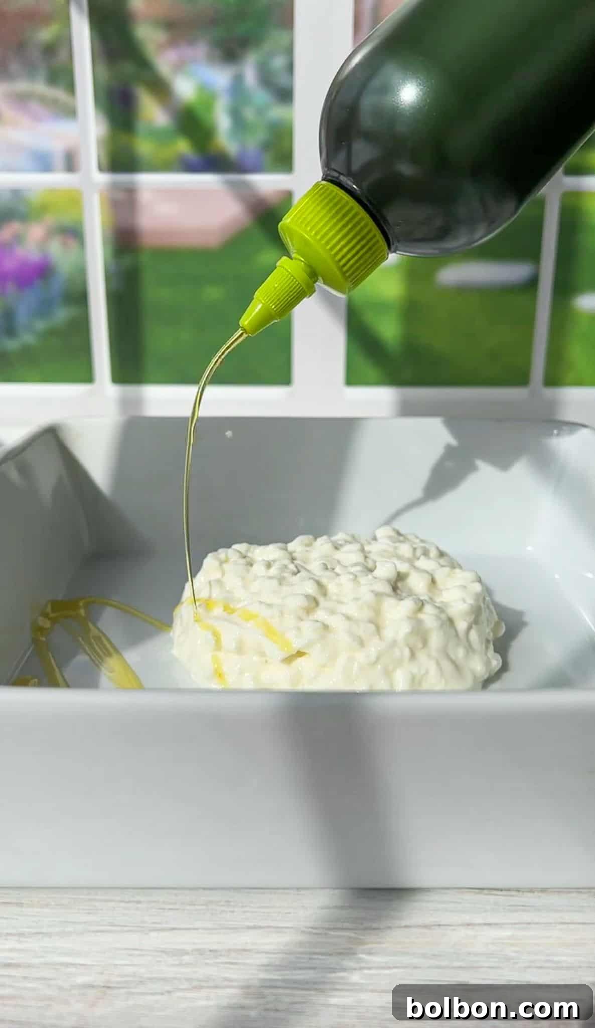 A black bottle with a green nozzle pours oil over a round piece of burrata cheese in a white square dish. The background features blurred garden scenery visible through a window.