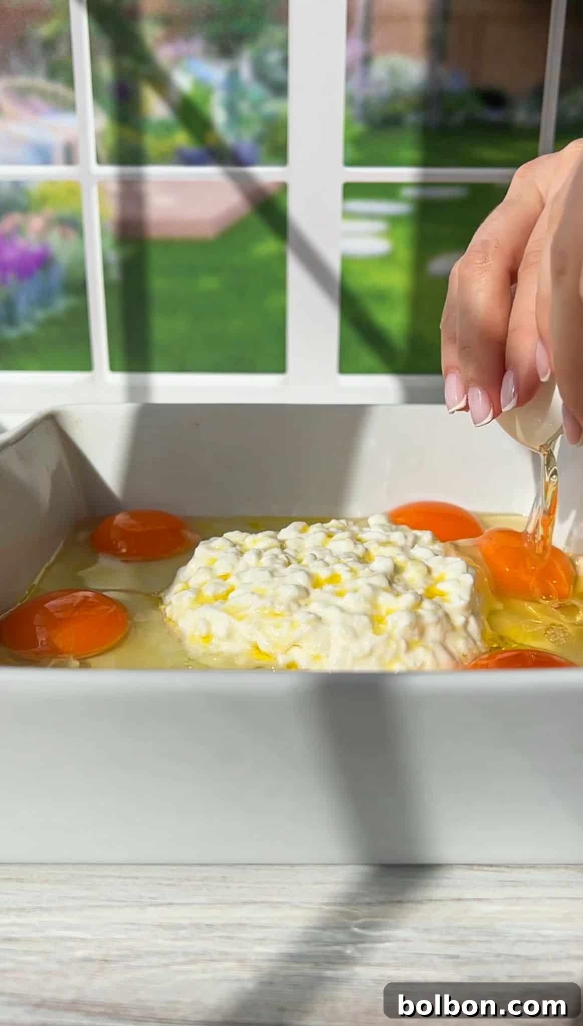 A hand cracking an egg into a white baking dish containing several eggs and a pile of cottage cheese. The background shows a window with a view of a garden.