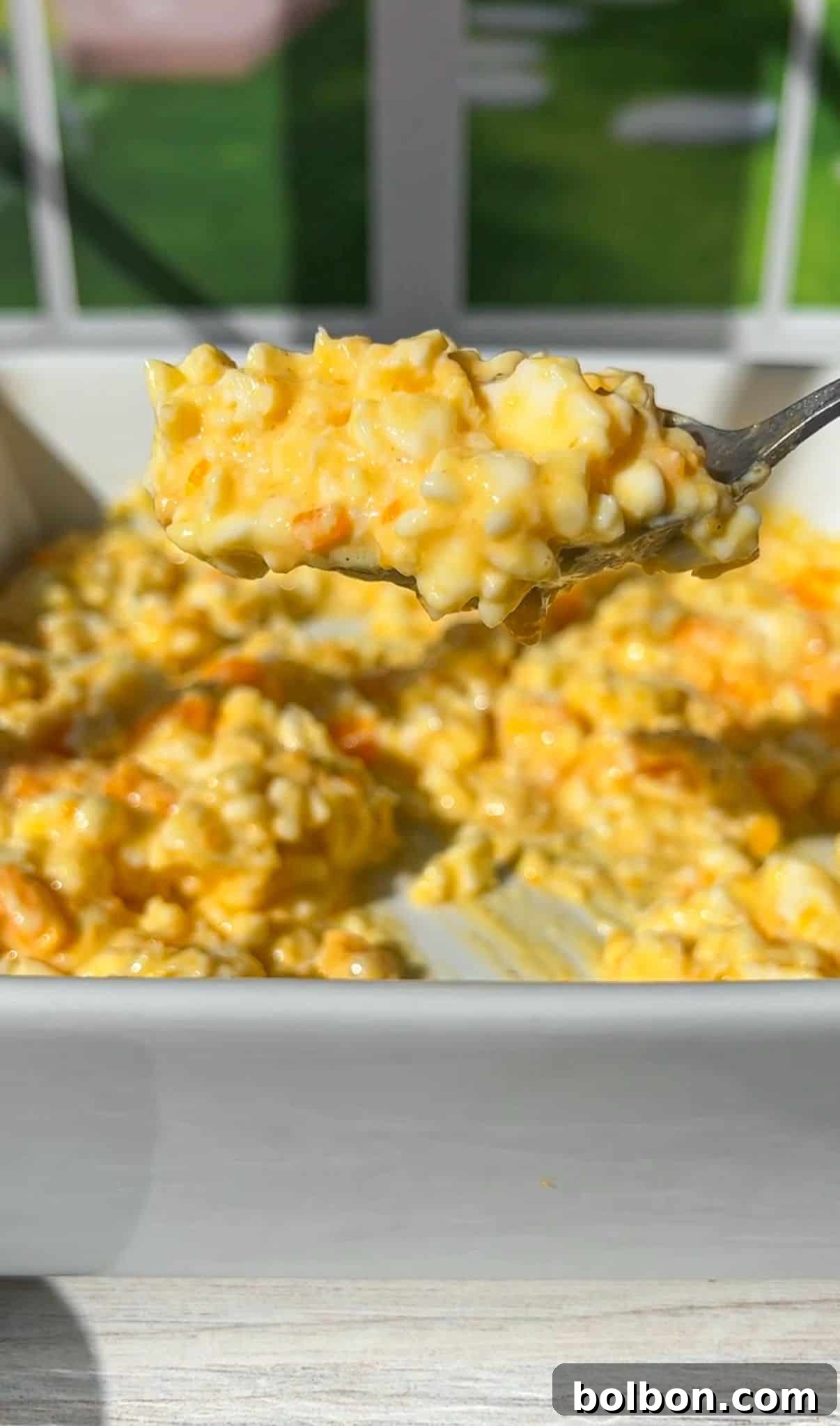 A spoonful of cottage cheese eggs is held above a white baking dish. The dish is filled with creamy, golden casserole, with visible chunks of potatoes and melted cheese. A sunlit window is blurred in the background.