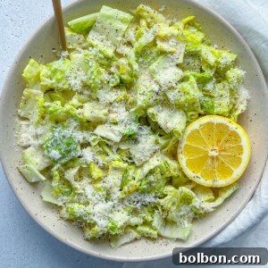 A bowl of chopped romaine lettuce forms the base of this classic Caesar Salad, topped with grated cheese and cracked pepper, garnished with a halved lemon, set on a light surface with a wooden fork and white napkin nearby.