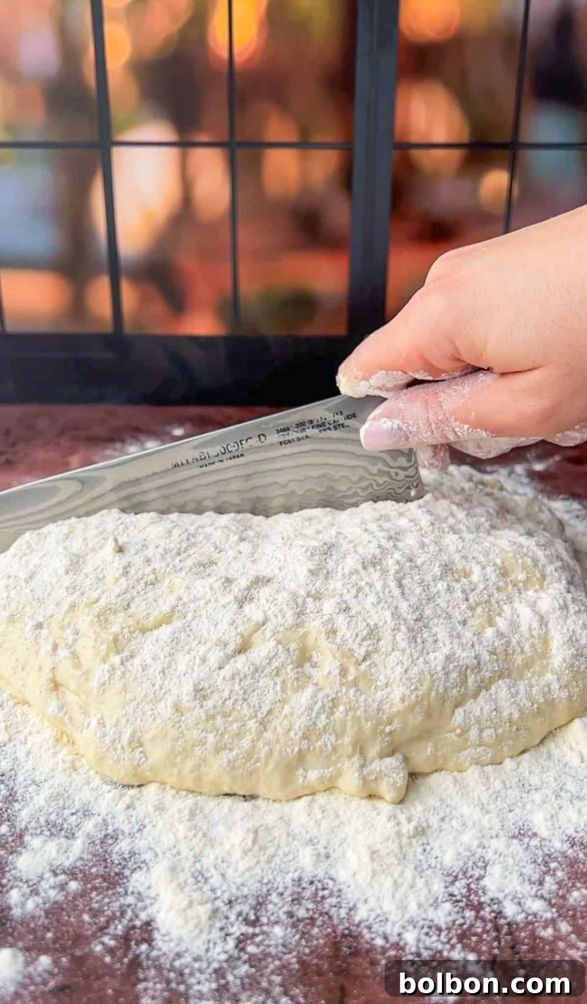 A close-up of a hand using a serrated knife to cut a floured dough on a brown surface. The dough is covered in a layer of flour. There is a blurred background with warm lighting.