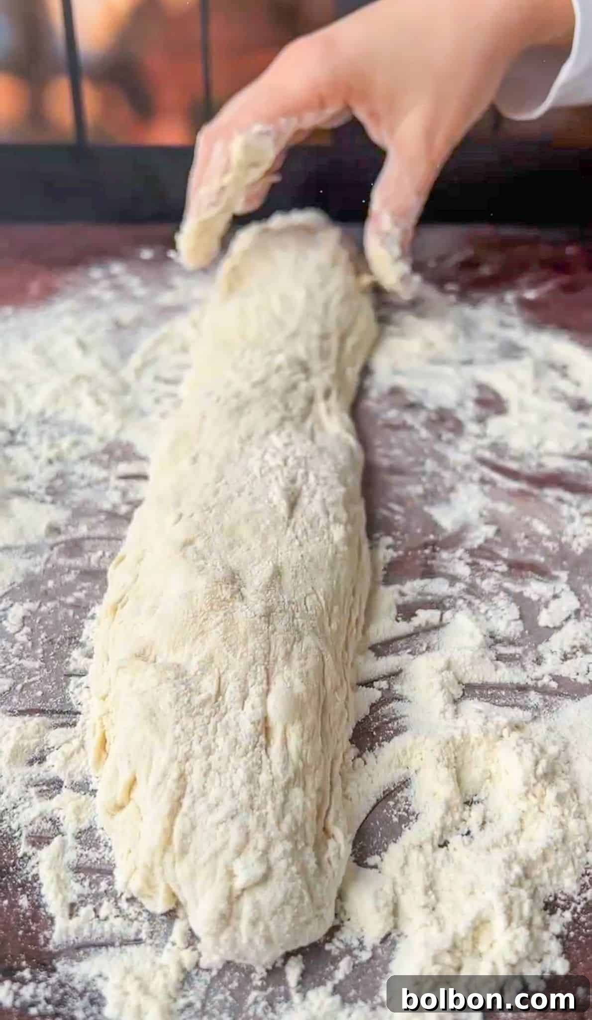 A person shaping a long piece of bread dough on a floured surface, with one hand gently folding the dough. The dough is lightly dusted with flour, and the surface is covered in flour as well.