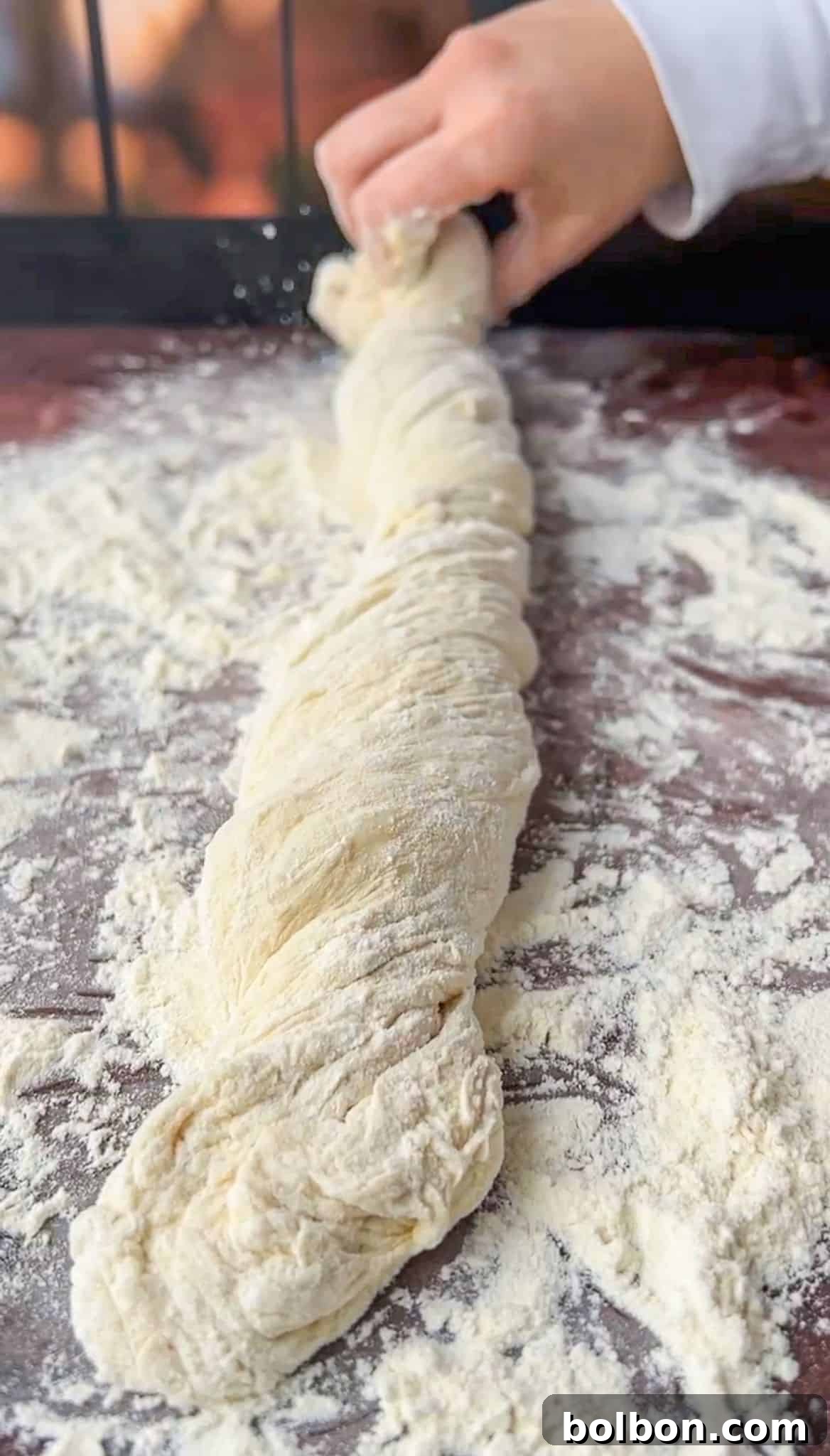 A hand is seen twisting a long, flour-dusted piece of dough on a floured wooden surface, preparing it for baking. The dough appears soft and fresh, with visible folds and texture.