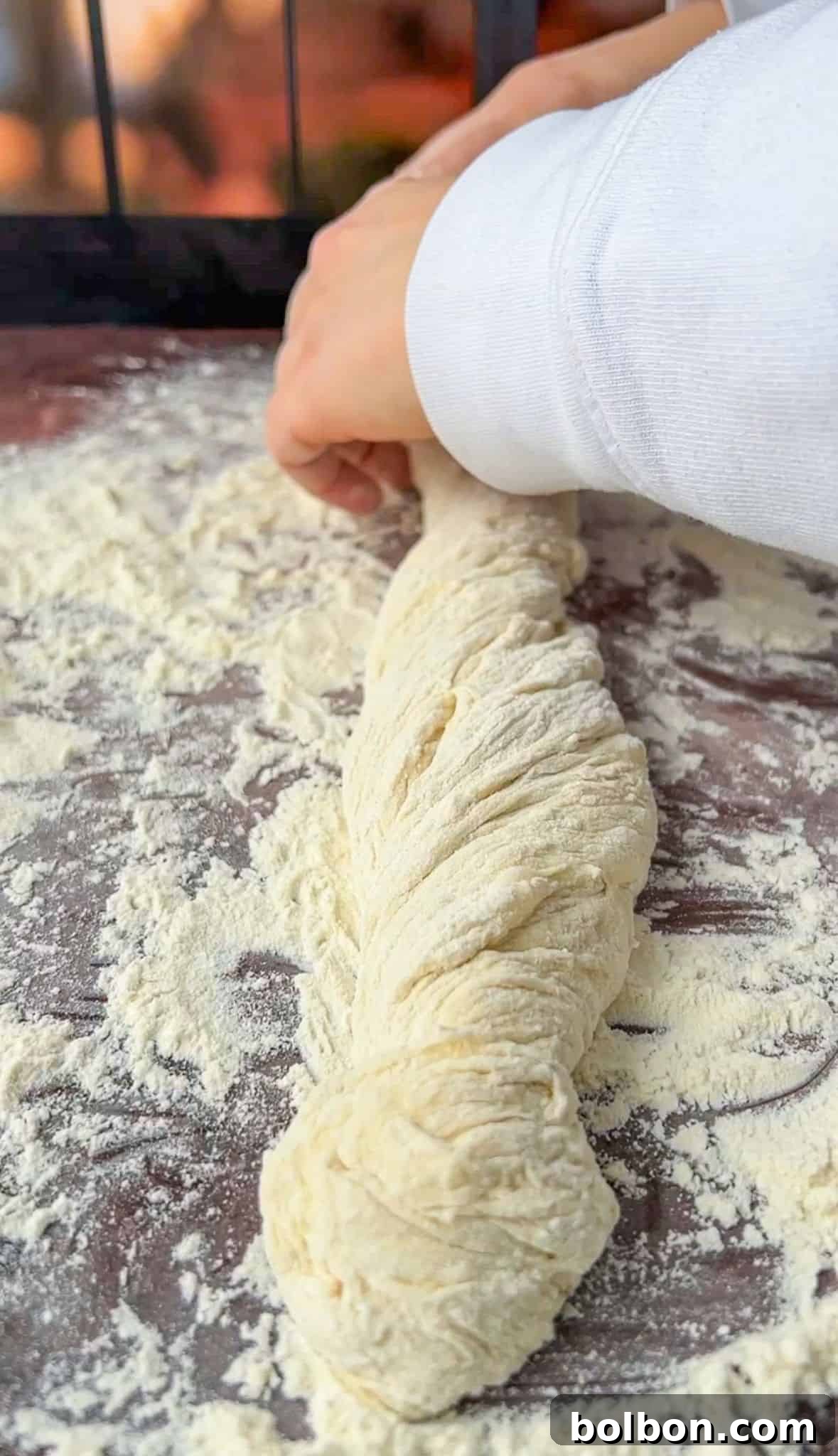 Person twisting a rope of dough on a floured surface, preparing bread or pastry. The dough is lightly dusted with flour, and the person's hand adjusts the twist.