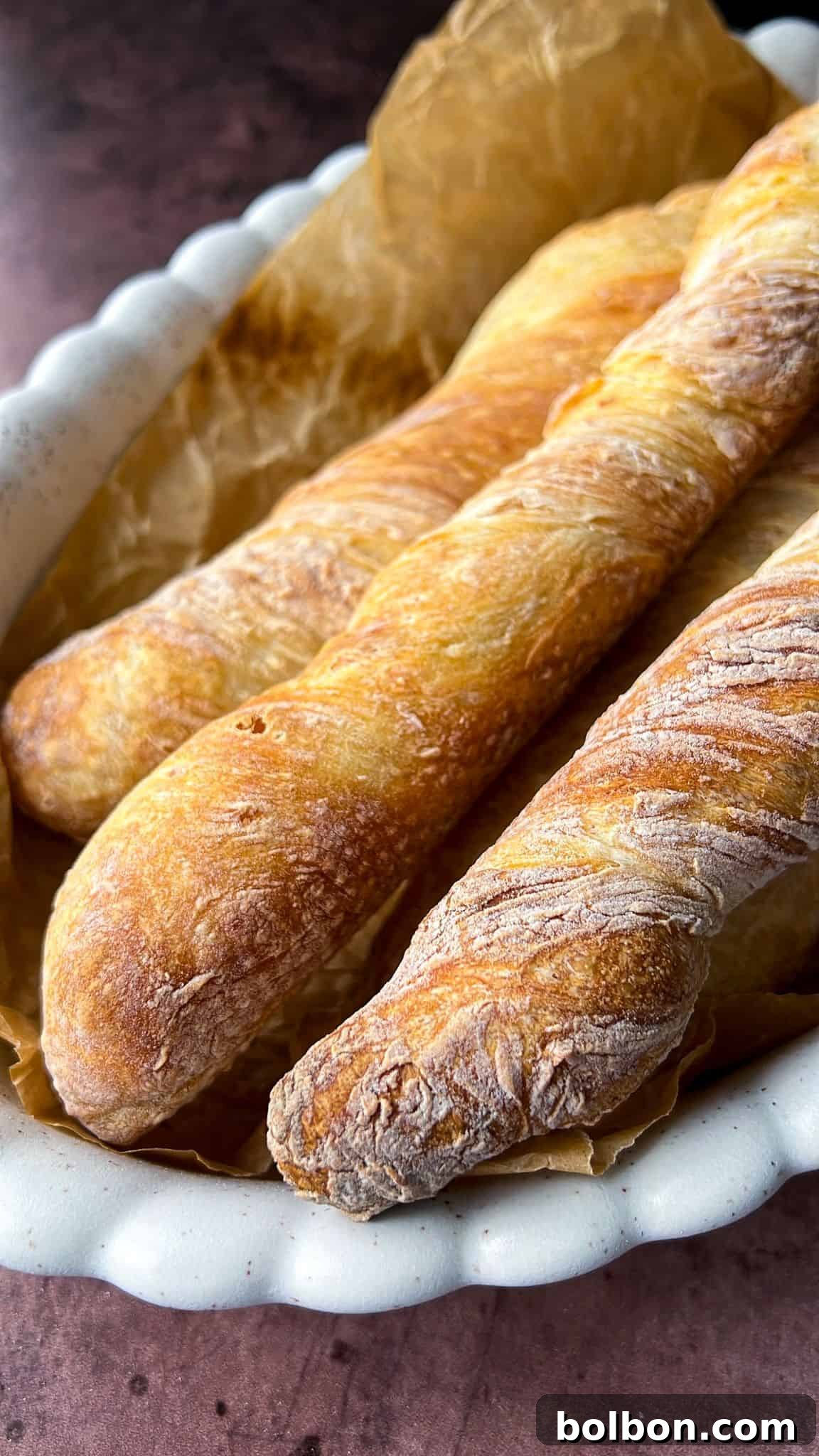 Three freshly baked baguettes on parchment paper in a white ceramic tray. The baguettes have a golden-brown crust and are placed on a rustic surface.