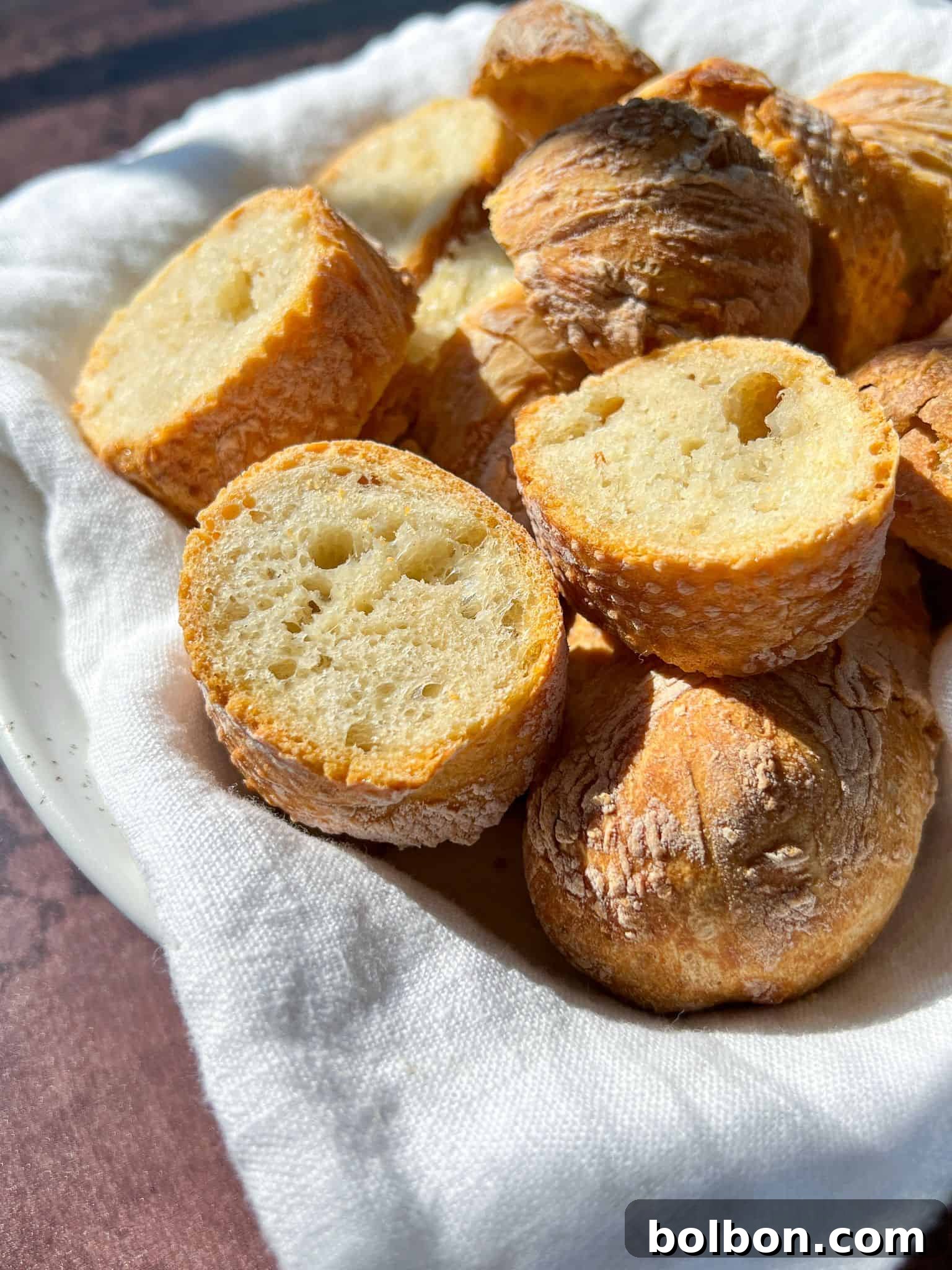 A white bowl lined with a cloth holds several pieces of sliced baguette. The bread has a golden crust and a soft, airy interior. Sunlight highlights the texture, casting gentle shadows on the surface.