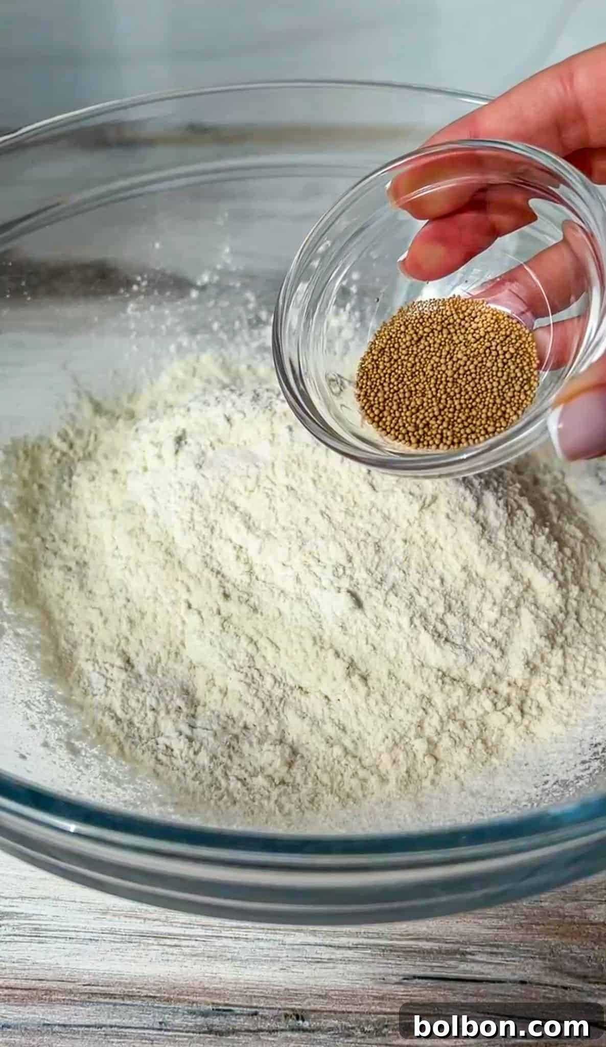 A clear glass bowl containing flour, with a hand holding a smaller glass bowl of brown mustard seeds above it. The scene is set on a light wooden surface.