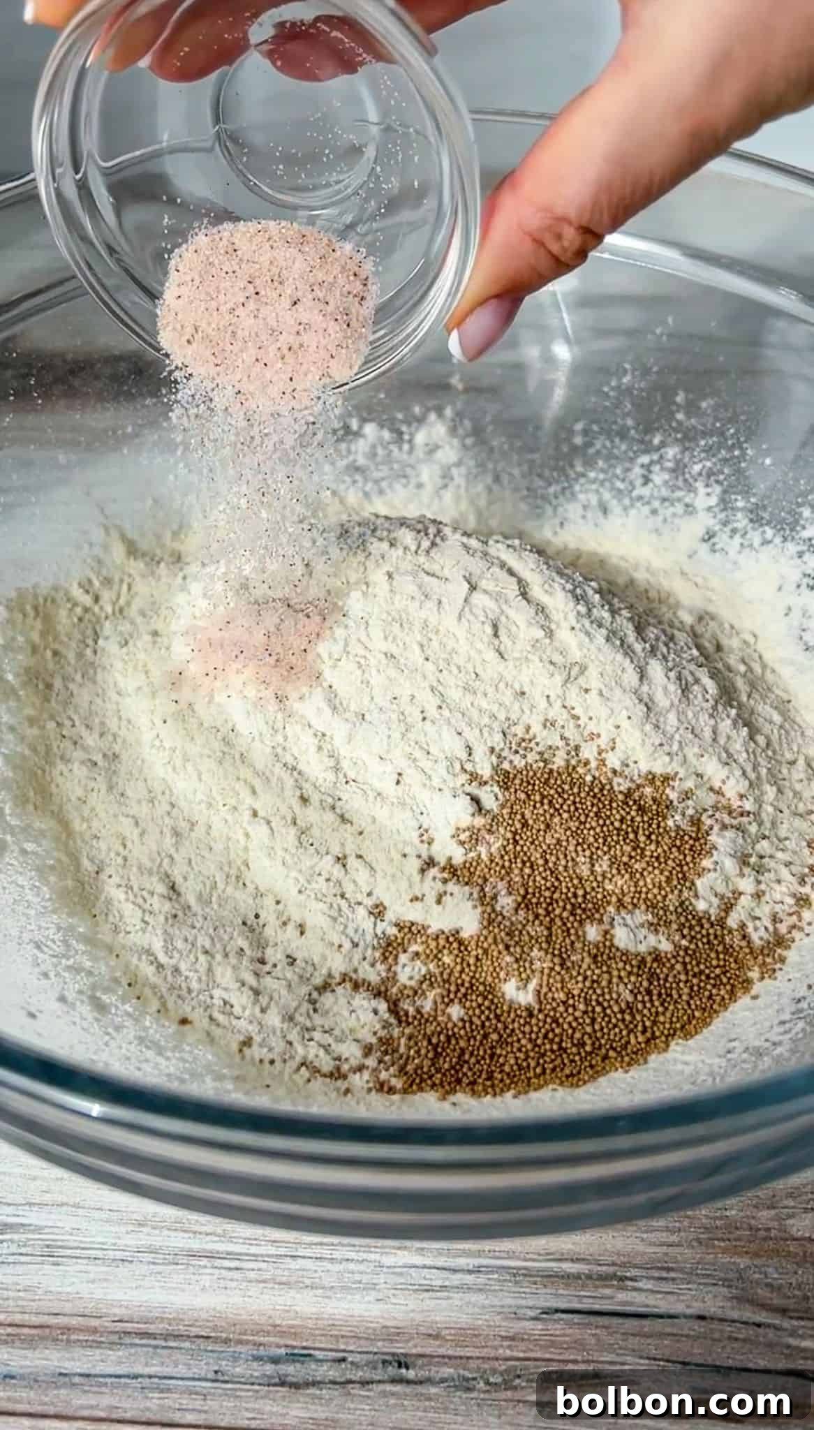 A hand pours pink salt and other seasonings from a small glass bowl into a large clear bowl containing flour and small brown seeds, likely yeast, on a wooden surface.
