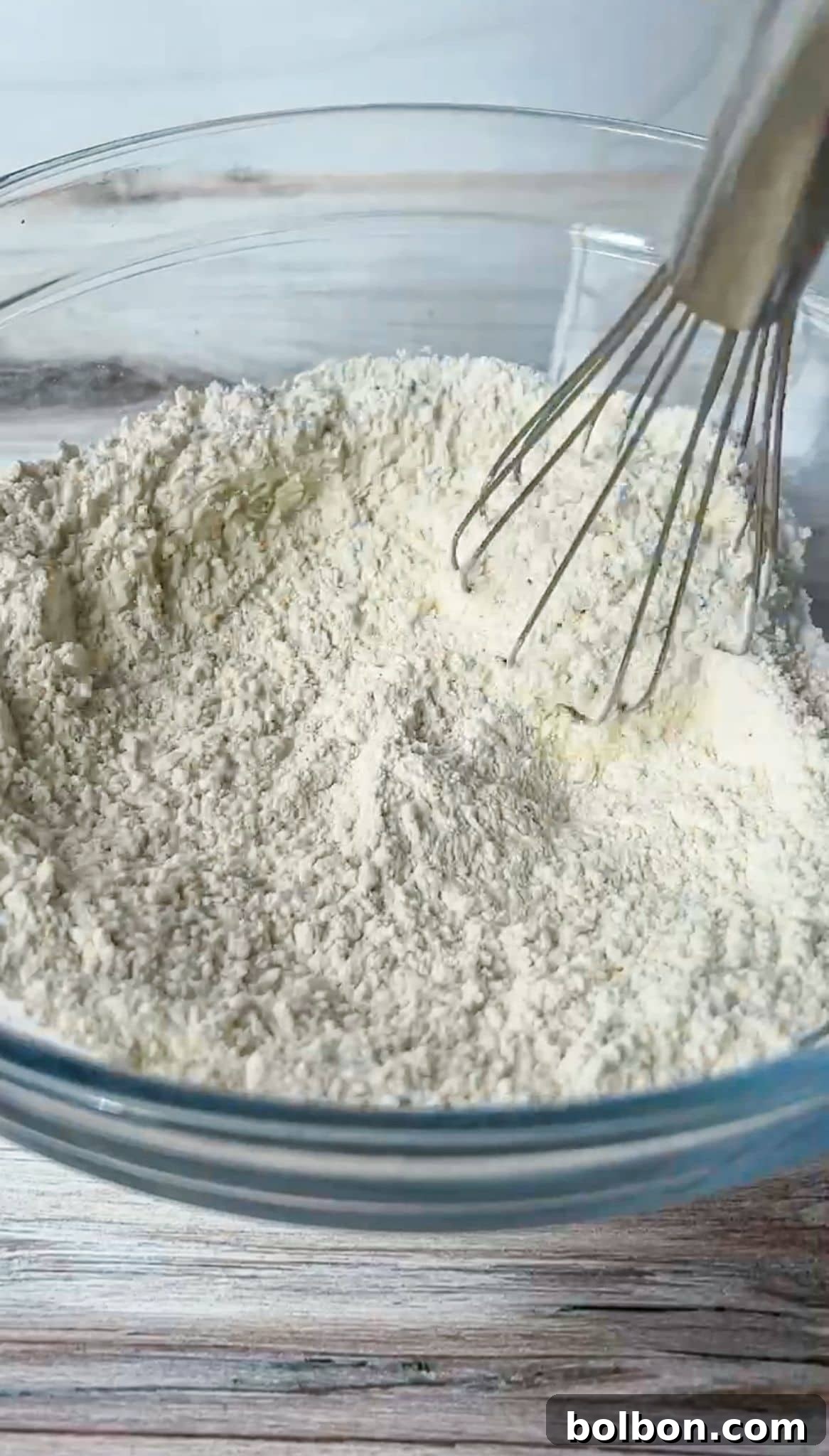 A whisk blending flour in a clear glass bowl on a wooden surface. The flour appears finely sifted and pale in color.