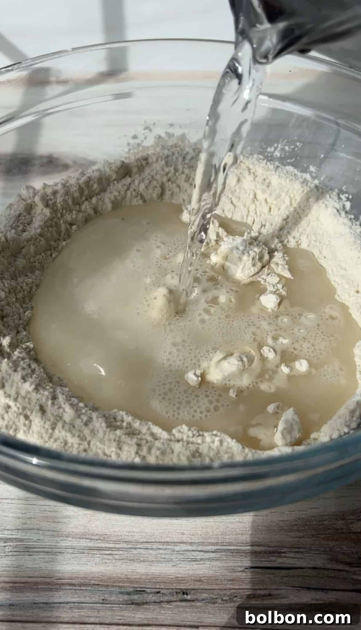Water being poured into a glass bowl filled with flour, creating a well in the center. The scene is set on a light wooden surface, capturing the moment before mixing ingredients.