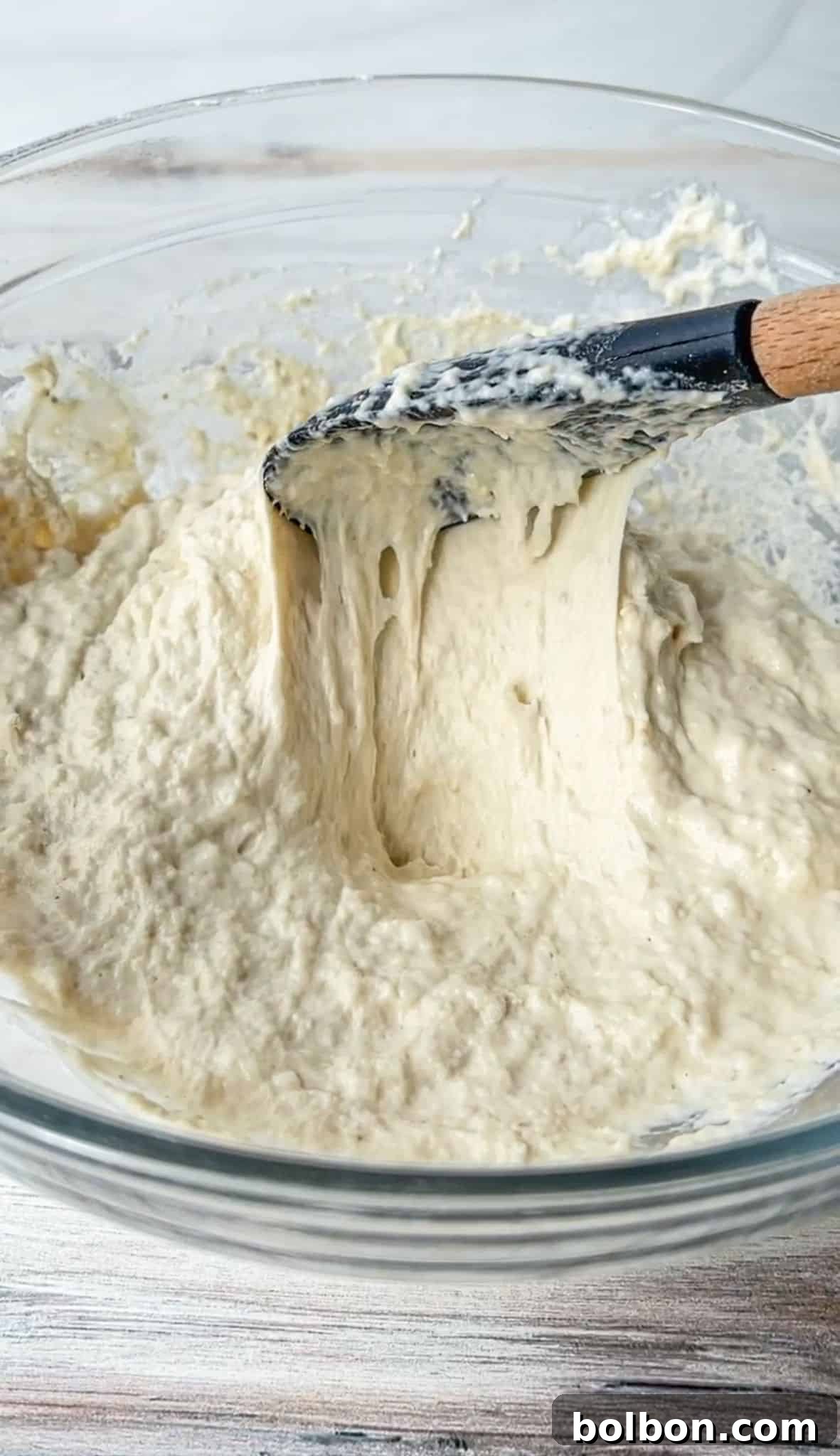 A wooden-handled spatula is mixing a thick, creamy batter in a clear glass bowl. The batter appears smooth and slightly sticky, with a pale color. The bowl is placed on a light wooden surface.