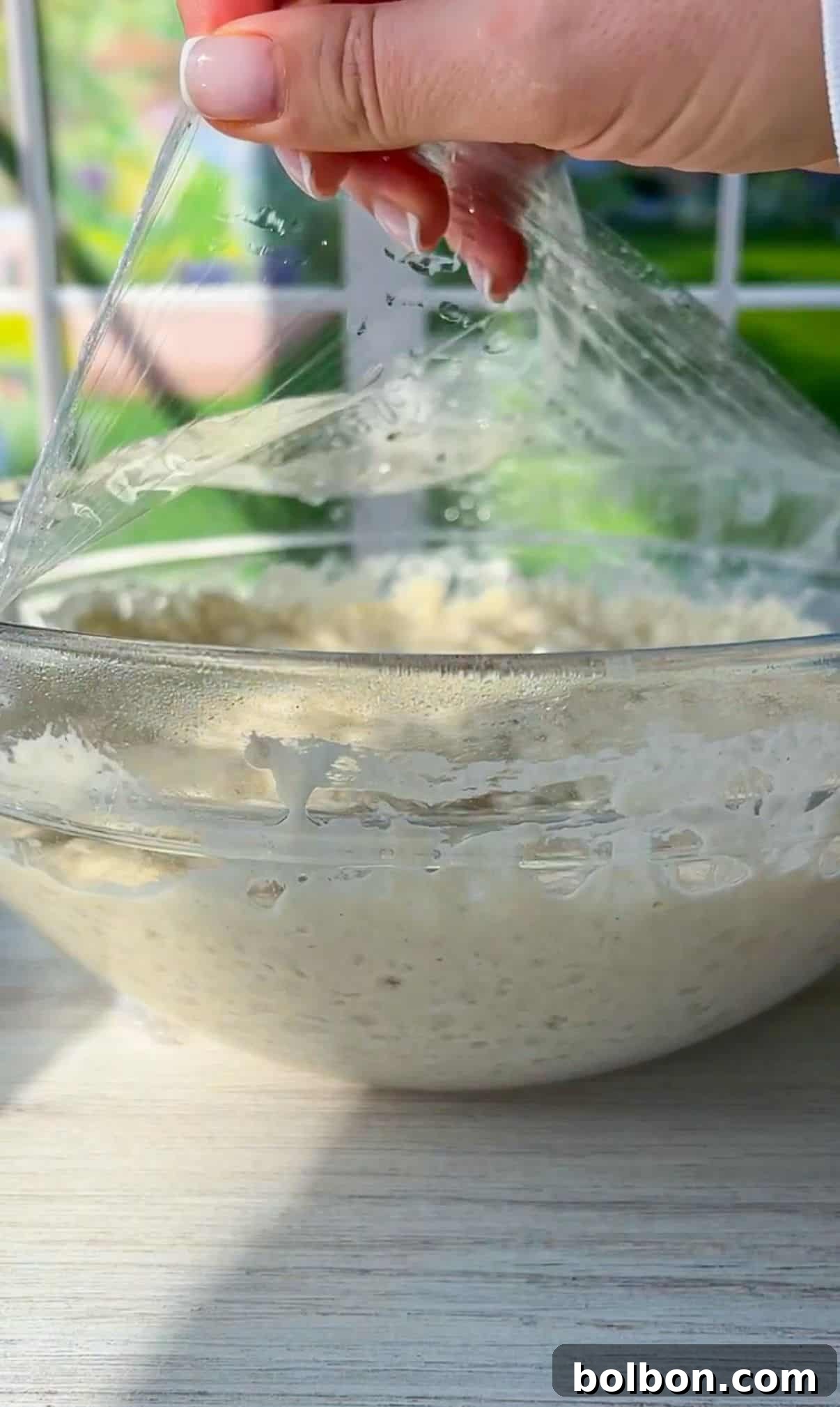 A hand is lifting plastic wrap off a clear glass bowl containing dough. The sunlight is illuminating the dough, highlighting its texture. The background shows a window with a blurred view of greenery outside.