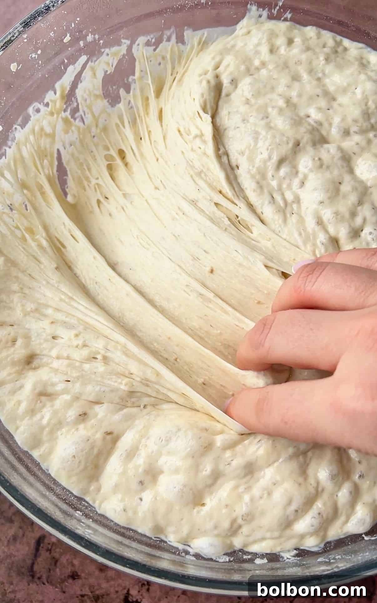 A hand stretches a portion of sticky, bubbly bread dough in a glass bowl. The dough appears well-kneaded and slightly elastic, with visible air pockets indicating fermentation and proofing. The background is a textured brown surface.