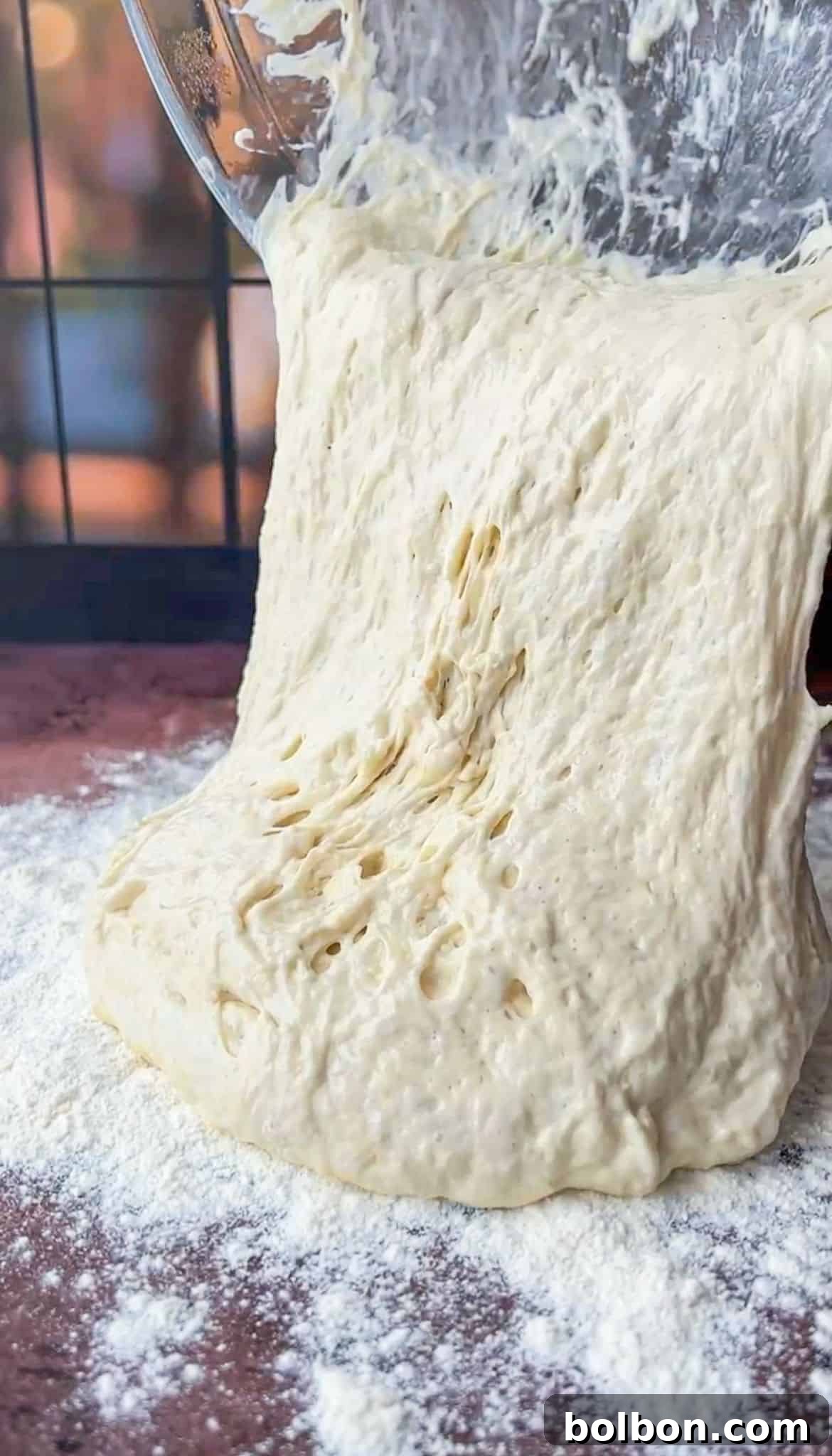 A close-up of sticky dough being poured from a bowl onto a floured surface. The dough has a light, airy texture, and the surface beneath is dusted with flour. A blurred background with hints of warm light is visible.