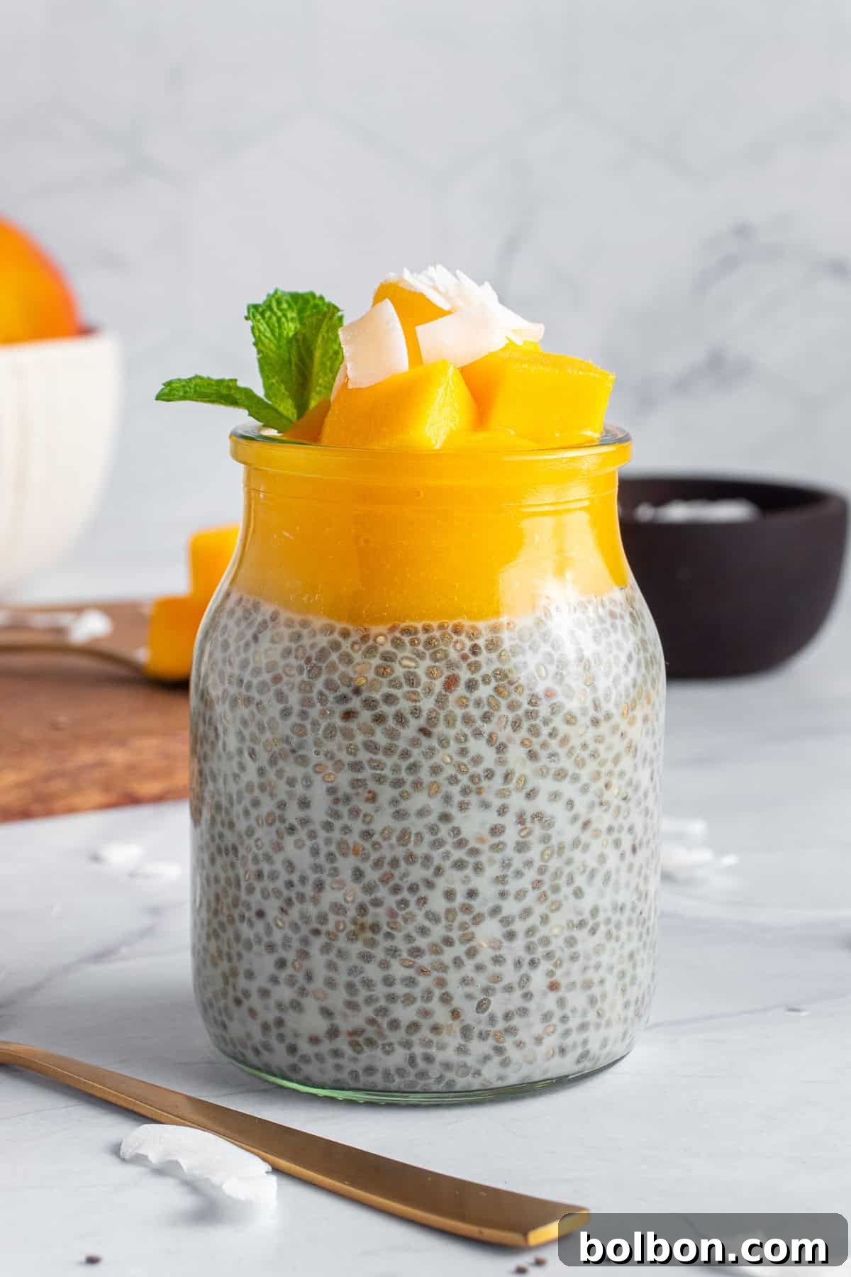 Tropical Mango Chia Delight 2 A jar of chia pudding topped with fresh mango chunks, coconut flakes, and a mint sprig, placed on a marble surface. A gold spoon lies nearby, with a blurred background of a bowl and a dark cup.