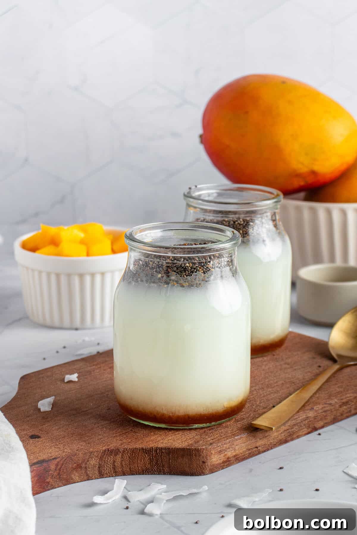 Tropical Mango Chia Delight 4 Two glass jars of a chia seed pudding mixture sit on a wooden board. Fresh mango chunks are in a small white bowl, with a whole mango nearby. A gold spoon rests on the board, and a light, hexagonal-tiled background completes the scene.