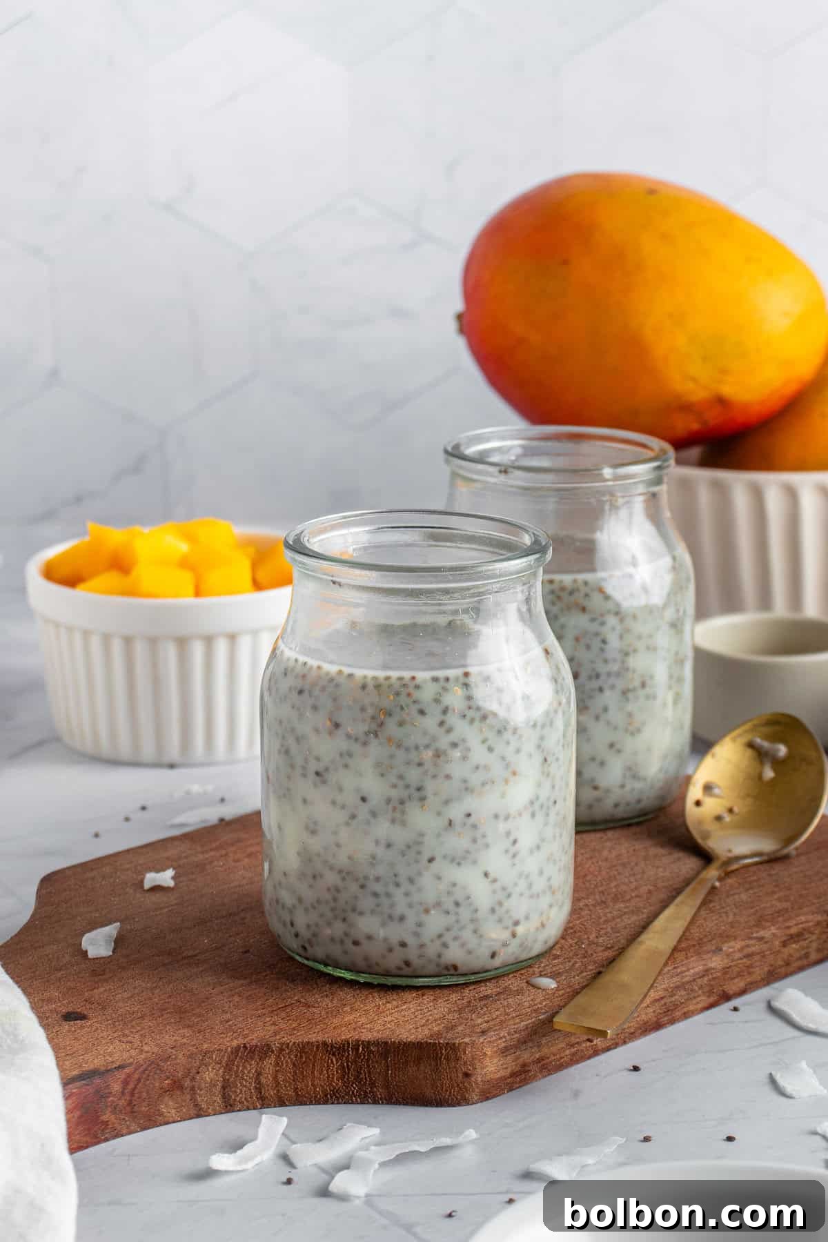 Tropical Mango Chia Delight 5 Two glass jars filled with the pudding mixture on a wooden board. A golden spoon rests beside them. In the background, a bowl of diced mango and two whole mangoes are visible. The setting is a marble surface with a subtle hexagonal pattern wall.