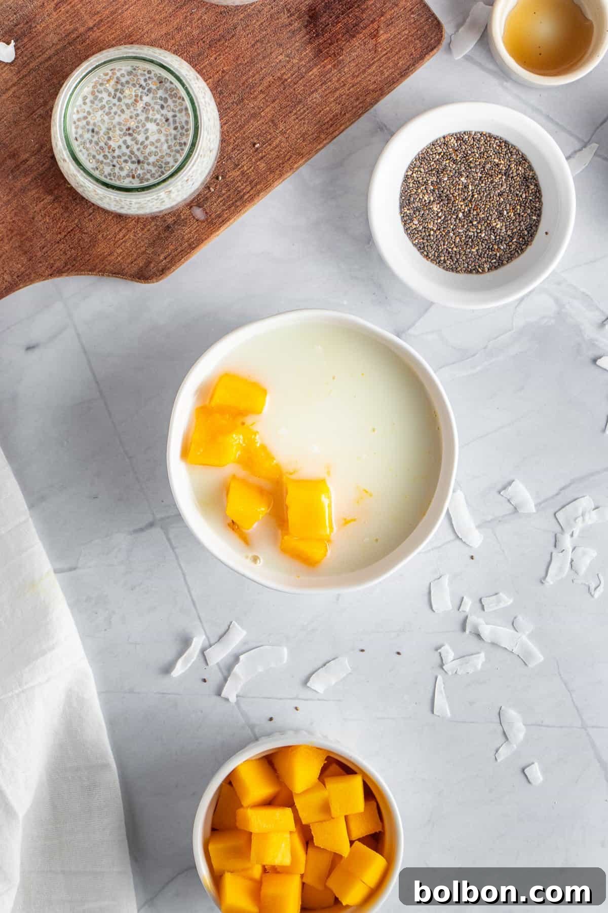 Tropical Mango Chia Delight 6 A white bowl of yogurt topped with mango chunks on a marble surface. Nearby are small bowls containing chia seeds, and diced mango. Coconut flakes and a wooden board are also visible.
