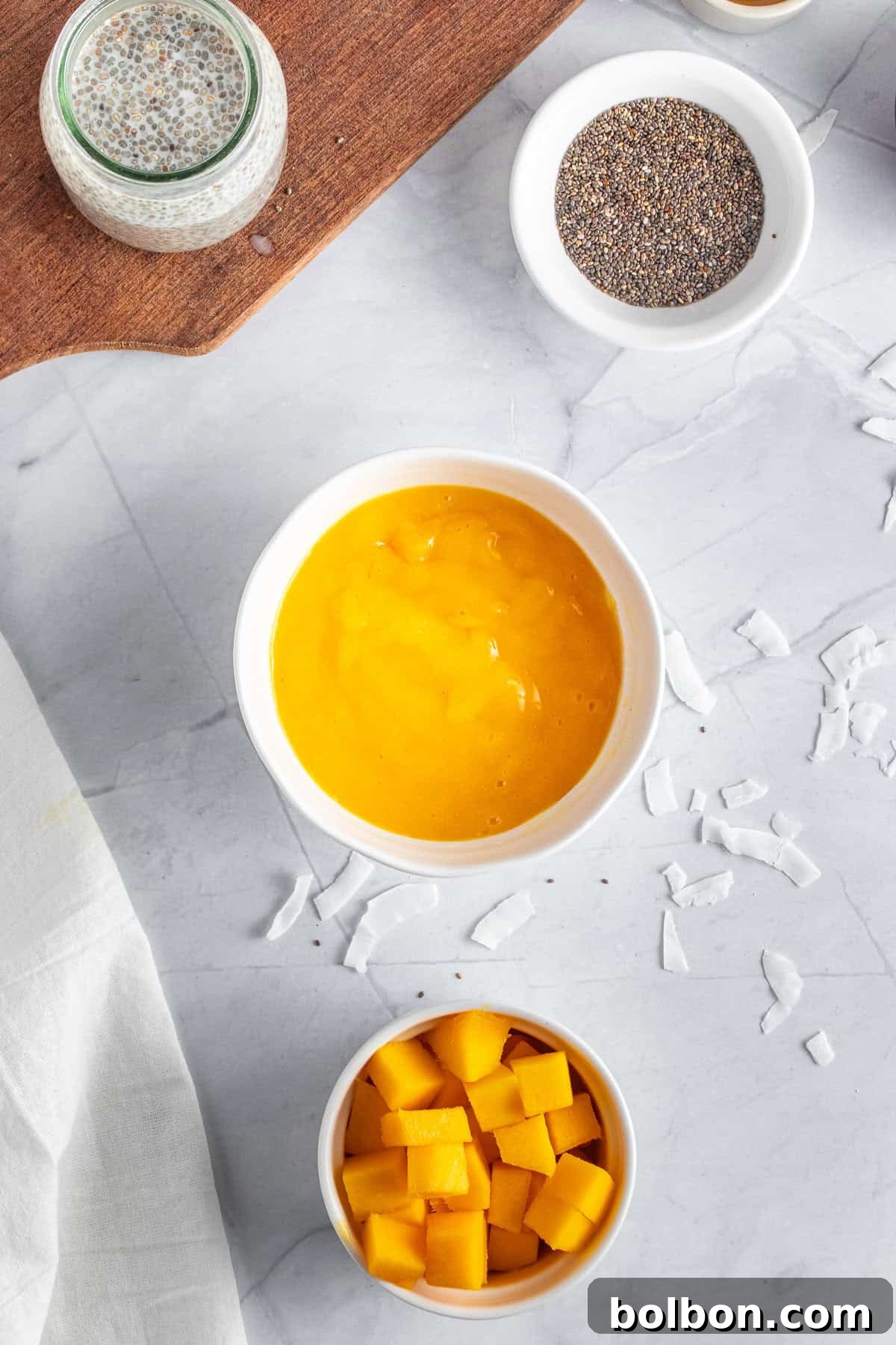 Tropical Mango Chia Delight 7 Overhead view of a smoothie bowl with bright yellow mango puree in a white bowl, surrounded by bowls of chia seeds, diced mango, and coconut flakes on a marble countertop. A cutting board with ingredients is partly visible.