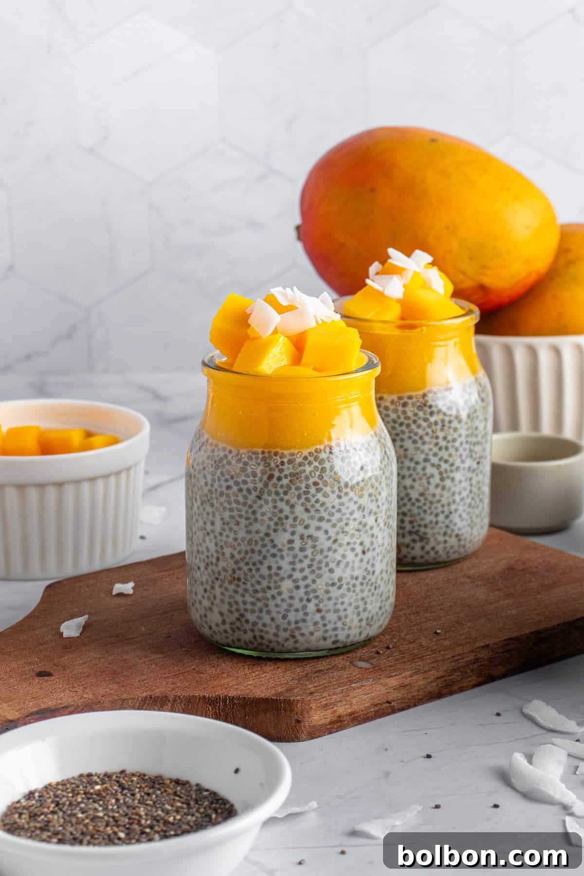 Tropical Mango Chia Delight 8 Two jars topped with diced mango and coconut flakes sit on a wooden board. A bowl of chia seeds and mango pieces are nearby. Whole mangoes are in the background against a light-colored geometric tile wall.