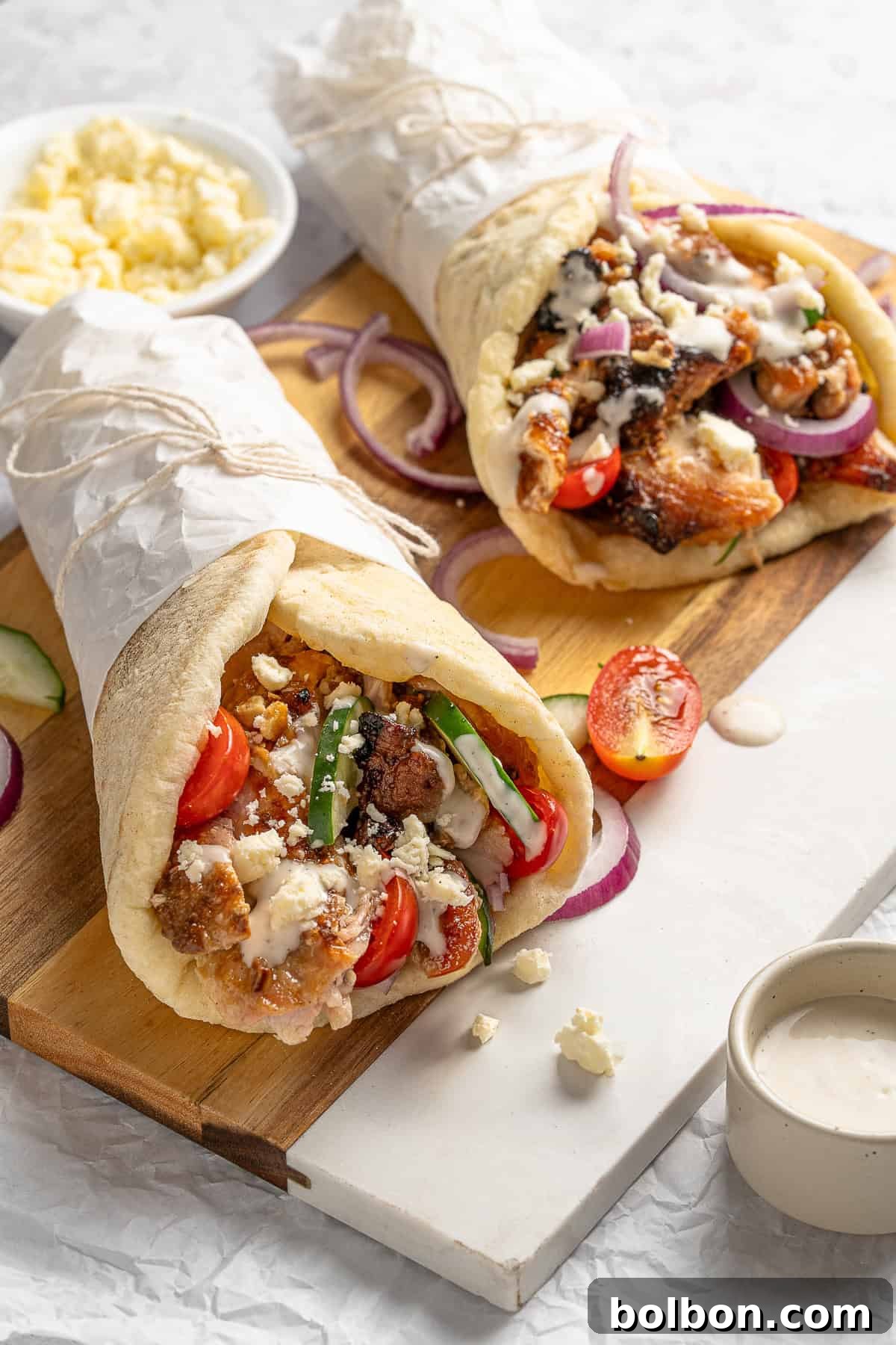 Effortless Greek Chicken Pitas 2 Two grilled pita wraps filled with chicken, tomatoes, cucumbers, red onions, and feta cheese drizzled with a creamy sauce, placed on a wooden board. A bowl of crumbled feta and a small dish of sauce are next to the wraps.