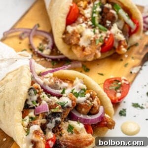 Two gyros filled with grilled meat, fresh tomatoes, red onion slices, crumbled feta, and herbs are served on a wooden board. A creamy sauce is drizzled on top, and the gyros are wrapped in pita bread.