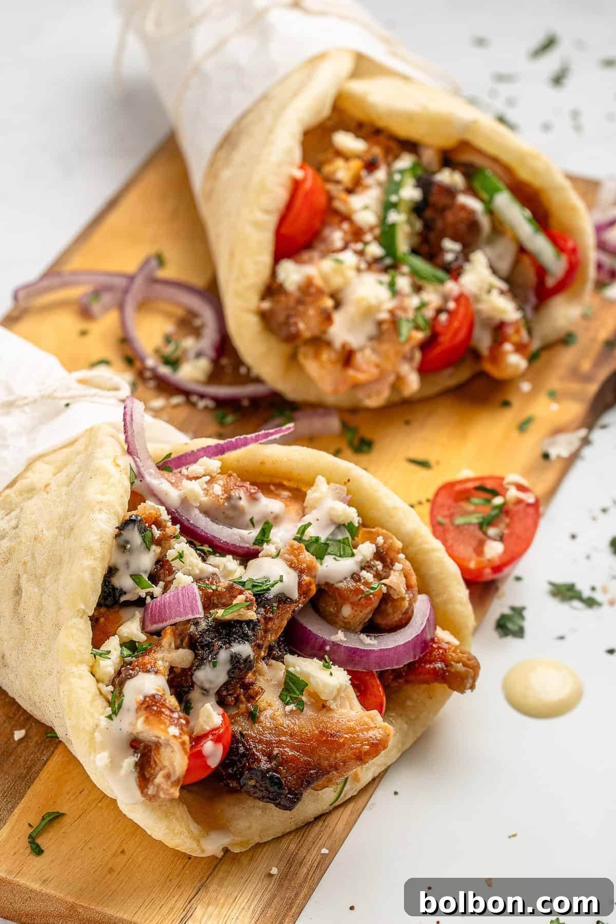 Effortless Greek Chicken Pitas 3 Two gyros filled with grilled meat, fresh tomatoes, red onion slices, crumbled feta, and herbs are served on a wooden board. A creamy sauce is drizzled on top, and the gyros are wrapped in pita bread.