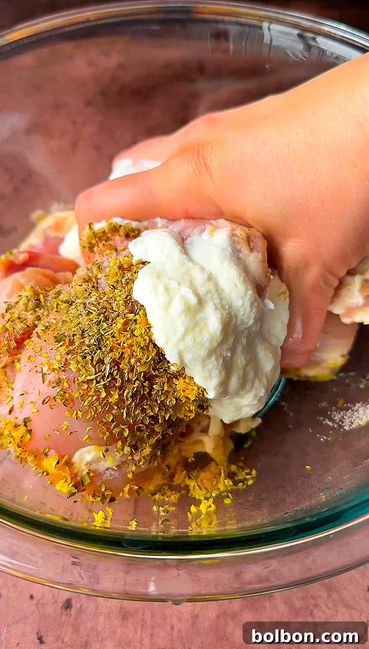 Effortless Greek Chicken Pitas 5 Hand mixing raw chicken pieces with a marinade of yogurt, herbs, and spices in a glass bowl.