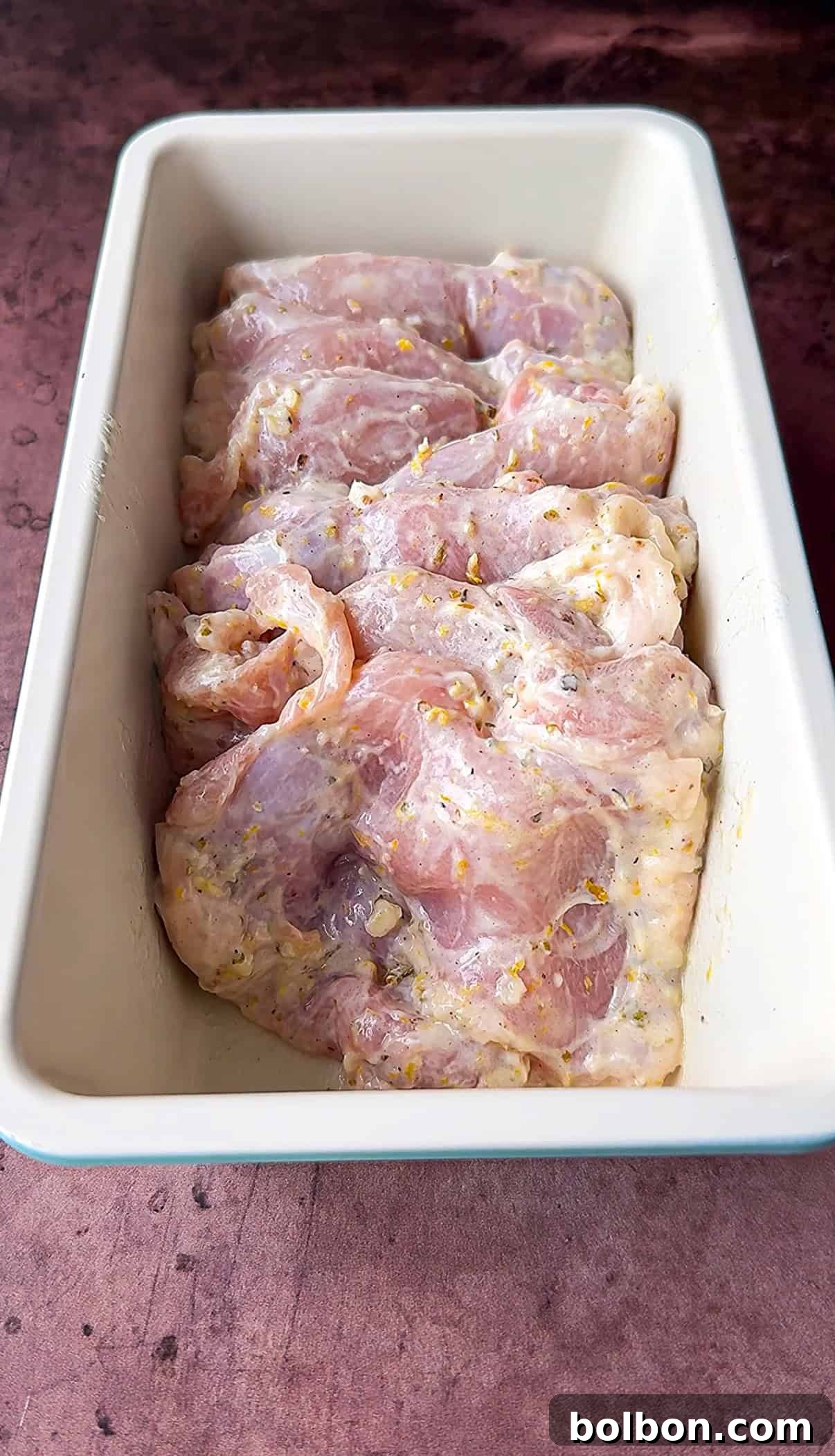 Effortless Greek Chicken Pitas 6 Raw chicken marinating in a rectangular pan, covered with a mixture of herbs and spices. The marinade appears to contain visible pieces of garlic and other seasonings. The pan is placed on a dark, textured surface.
