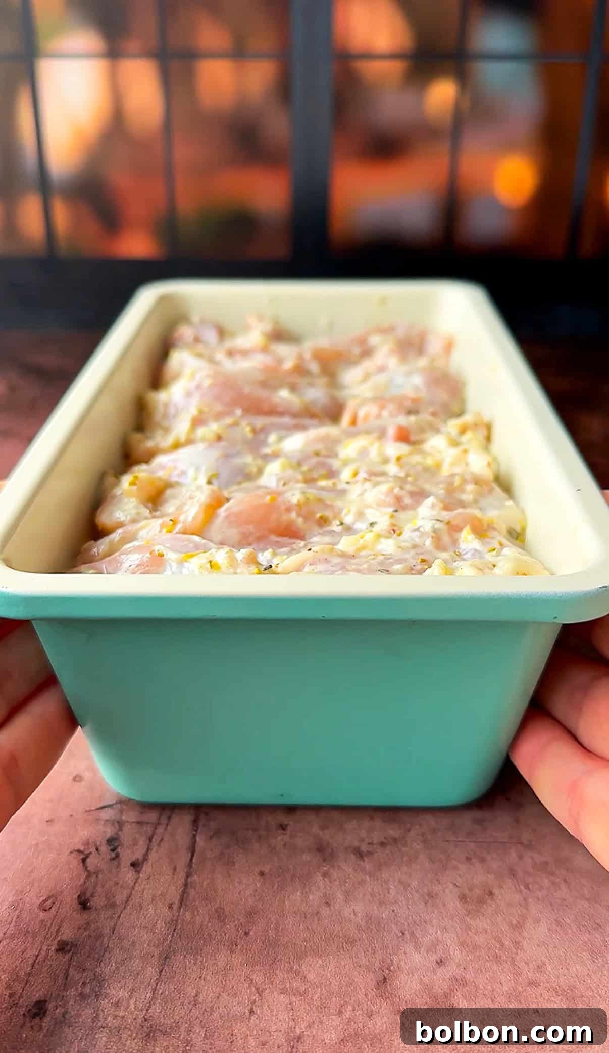 Effortless Greek Chicken Pitas 7 Hands holding a light blue loaf pan filled with marinated thighs, ready to be cooked. The background shows a warm, softly lit kitchen.