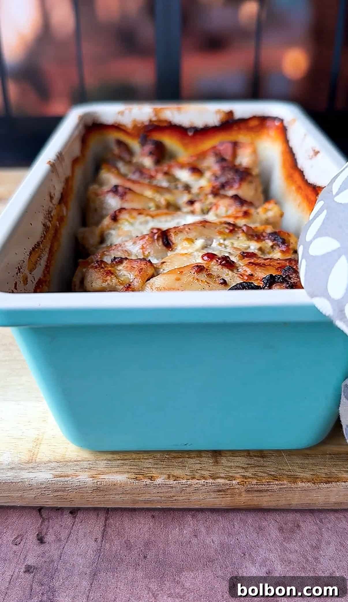 Effortless Greek Chicken Pitas 8 A turquoise rectangular baking dish containing golden-brown baked thighs with visible crispy edges. The dish is resting on a wooden surface, partially covered by a gray cloth with white leaf patterns.