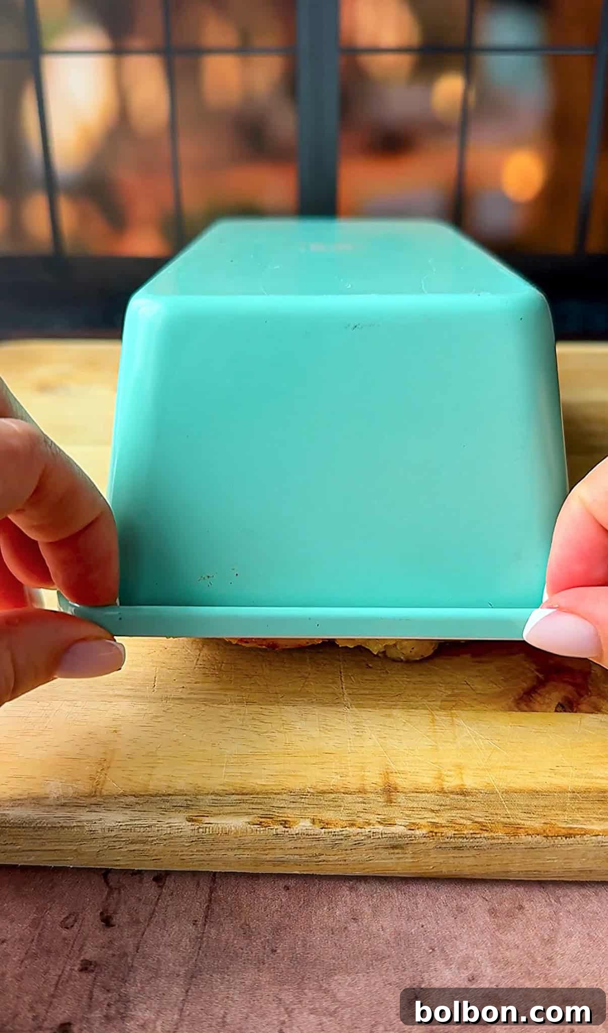 Effortless Greek Chicken Pitas 9 Two hands lift the edge of an upside-down, teal rectangular container placed on a wooden cutting board. The background features a black window frame with blurred outdoor scenery.