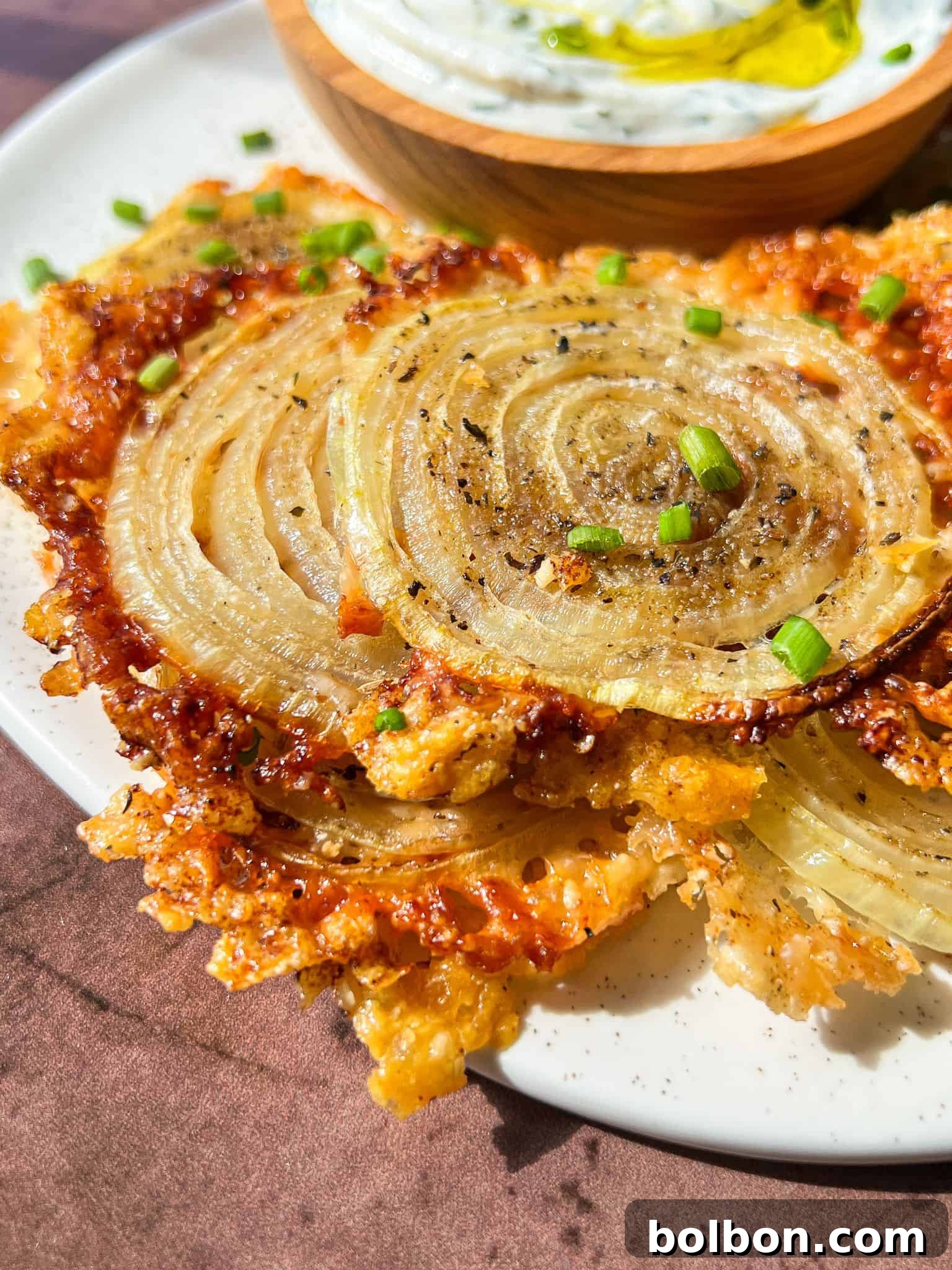 Trending Onion Ring Crisps 2 Crispy, golden onion ring chips and chopped chives placed on a white plate. In the background, a small wooden bowl contains a creamy dip with a drizzle of olive oil.