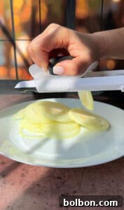 A hand is slicing a white onion using a handheld slicer over a white plate. Sunlight creates soft shadows on the wooden surface, enhancing the kitchen setting's warm ambiance.
