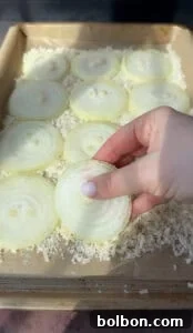 A hand holding a slice of raw onion over a tray filled with multiple onion slices and grated Parmesan cheese. The scene is well-lit, emphasizing the layers and texture of the onions.