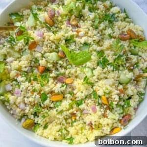 A white bowl filled with fresh Jennifer Aniston Couscous Salad, featuring chopped cucumber, red onion, herbs, and garnished with pistachios and mint leaves. The bowl is set on a white surface.