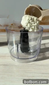 A measuring cup pours cottage cheese into a clear food processor bowl. The kitchen has a light wooden counter, and blurred kitchenware is visible in the background. Sunlight highlights the texture of the cheese.