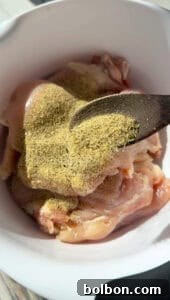 Raw chicken pieces in a white bowl are being sprinkled with a green and yellow seasoning mix from a spoon. The image is close-up, focusing on the seasoning process.