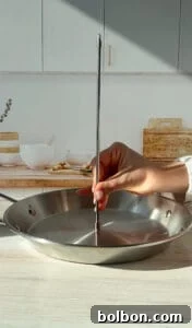 A hand holds a skewer or probe vertically in a stainless steel pan filled with water on a kitchen counter. In the background, kitchen items like bowls, a cutting board, and dried plants are visible.