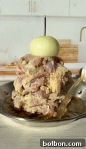 A pile of marinated raw meat stacked on a vertical skewer in a pan, topped with a whole onion. Liquid is being poured into the pan. The background shows a kitchen counter with a cutting board and faucet.