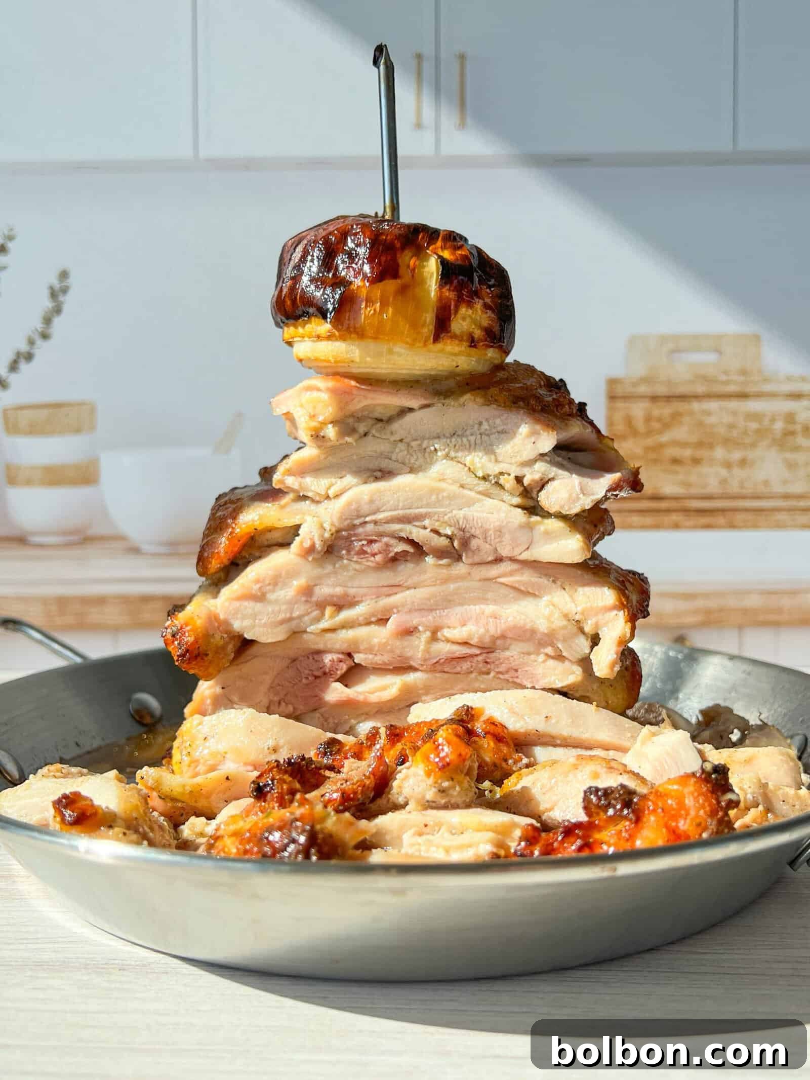 High Protein Chicken Gyro. A towering stack of sliced, roasted chicken pieces is skewered on a vertical rod in a pan. A roasted onion tops the stack. The setting includes a bright kitchen with a wooden cutting board and decorative jars in the background.