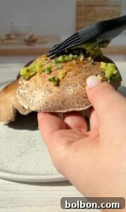 A hand holds a mushroom on a speckled plate while a black brush spreads a chopped green mixture on top. Sunlight creates a warm atmosphere in the kitchen.